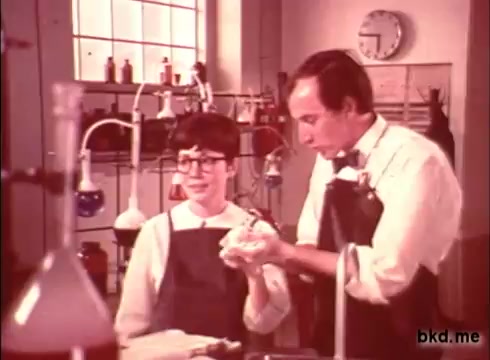 The 16mm still from the 1970s Camay Soap commercial features two individuals in a laboratory setting. One is a young boy wearing glasses, and the other is an adult man, both dressed in lab aprons. They are engaged in a demonstration, possibly involving soap or a related product, with various colorful laboratory equipment in the background. The atmosphere suggests an educational or promotional theme, typical of commercial filmmaking from that era.