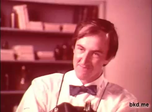 The still from the 1970s Camay Soap commercial features a man wearing a bow tie and an apron, smiling at the camera. The background shows shelves with various items, suggesting a domestic or grooming setting. The color tone appears warm and slightly faded, typical of 16mm film from that era.