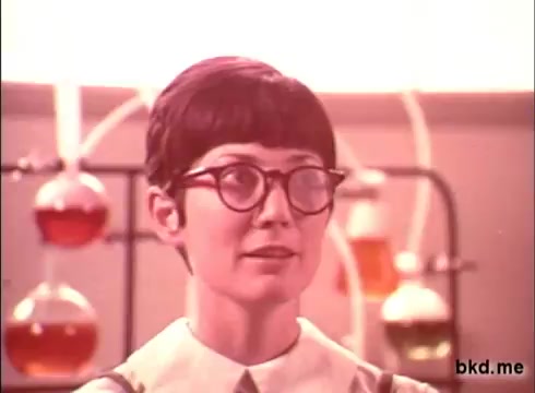 The still features a woman with short hair and glasses, set against a backdrop of laboratory glassware filled with colorful liquids. She appears to be explaining or demonstrating something, likely related to the Camay soap product. The overall aesthetic conveys a vintage 1970s style, highlighting a blend of science and beauty in the advertising context.