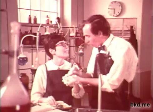 The still from the 1970s Camay soap commercial features two characters, a man and a woman, in a laboratory setting. The woman, wearing glasses and an apron, appears engaged in conversation with the man, who is also dressed in a white shirt and an apron. They are surrounded by various lab equipment and bottles, suggesting a scientific approach to the soap's formulation. The warm color tones typical of the era add to the nostalgic atmosphere of the scene.