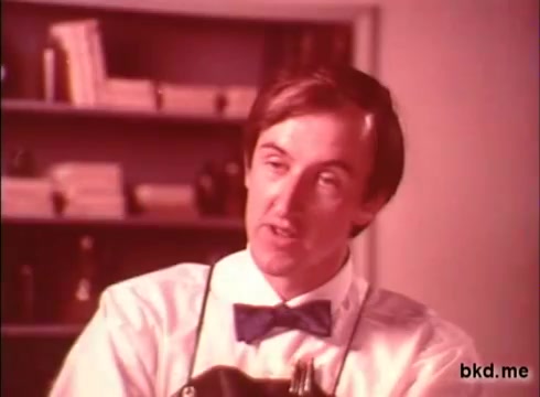 The still features a man in a bow tie and white shirt, likely portraying a barber or salesman, speaking directly to the camera. He has a slightly exaggerated expression, suggesting enthusiasm or persuasion. The background is softly focused, showing shelves with indistinct objects, evoking a vintage aesthetic typical of 1970s commercials. The overall color tone is warm, reflecting the era's visual style.
