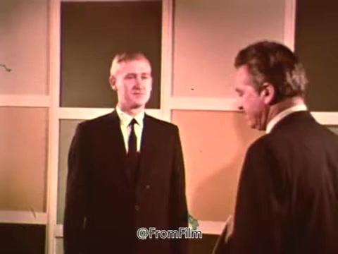 The still features two men in formal attire, likely representing different generations or roles, engaging in a conversation. The setting has a backdrop of muted, geometric shapes in soft colors, typical of the late 1960s design aesthetic. The overall tone conveys a professional atmosphere, suggesting a discussion related to G.I. Insurance and benefits for veterans, as indicated by the context of the PSA. The phrase 'For Information About - G.I. Insurance and other benefits check with your local V.A. Office' summarizes the purpose of the message, emphasizing accessibility to essential services for veterans.