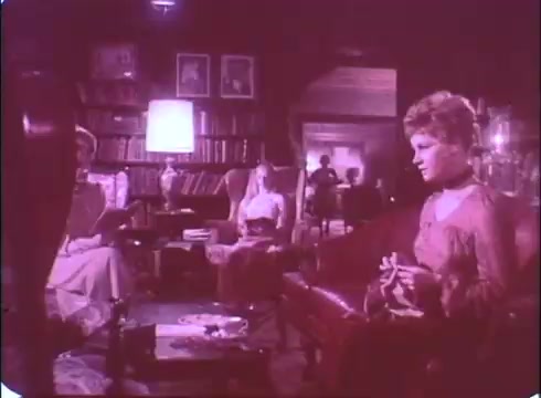 The still from the trailer of 'A Reflection of Fear' showcases a dimly lit interior, likely a living room or parlor, filled with vintage decor. Three women are seated, with one prominently in the foreground holding a cigarette and appearing contemplative. The atmosphere is tense and mysterious, with shadows adding to the eerie vibe. The background features bookshelves and indistinct figures that enhance the unsettling ambiance typical of a horror film. The faded colors typical of a 16mm print contribute to a nostalgic, unsettling look.