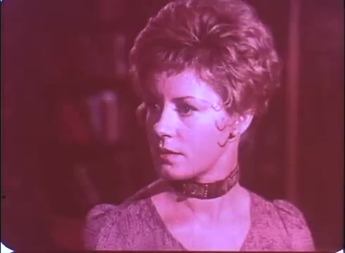The still features a young woman with short, styled hair, looking intently to the side. Her expression is dramatic, suggesting tension or concern, fitting for a horror flick. She's wearing a simple, elegant dress and a choker necklace, contributing to the vintage aesthetic of the 16mm print. The overall color tone is muted, with a reddish hue that adds to the unsettling atmosphere of the trailer.