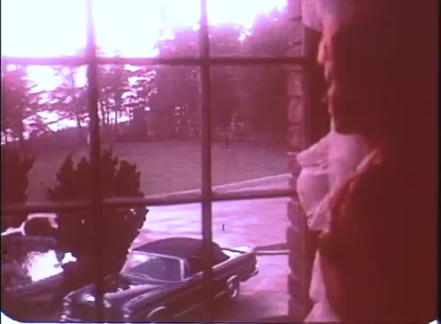 The still from 'A Reflection of Fear' trailer depicts a moody, atmospheric scene through a window. A figure, possibly a woman, stands gazing outside, shrouded in a sense of foreboding. The surrounding environment features a vintage car parked on a driveway, and the color tones suggest a faded quality typical of a 16mm print. The overall composition hints at a suspenseful narrative, reflective of classic horror film elements.
