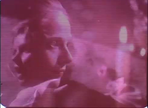 The still from the trailer of 'A Reflection of Fear' presents an eerie atmosphere typical of a horror film. It features a woman with a contemplative expression, illuminated by flickering candlelight. The color palette leans towards muted tones with a pinkish hue, creating a surreal, unsettling feel. The graininess of the 16mm print adds to its vintage charm, hinting at classic horror aesthetics while suggesting an ominous narrative involving suspense and intrigue.