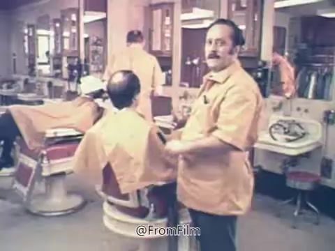 The still is from a Rolaids commercial, featuring a barbershop setting. A barber in an orange shirt stands next to a customer in a barber chair, while other customers are getting haircuts. The vibe is lighthearted and casual, emphasizing the playful interaction that ties into the iconic tagline, 'How do you spell relief? R-O-L-A-I-D-S.' This commercial highlighted Rolaids' role in providing relief, connecting it to Major League Baseball's Relief Man of the Year Award.