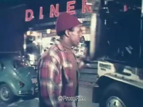 The still shows a scene outside a diner, featuring a man in a plaid shirt and a red beanie. The diner is brightly lit and has a vintage feel, indicative of the 1970s era. In the background, a green car is parked. This image relates to the iconic Rolaids commercial known for its tagline 'How do you spell relief?' which highlights the brand's association with providing relief from indigestion. The commercial was significant enough to lead to Rolaids sponsoring the Rolaids Relief Man of the Year Award in Major League Baseball.