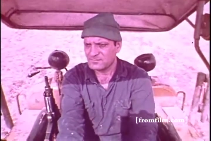 The still features a man driving a tractor, likely set in an agricultural or rural context. He has a focused expression, wearing a green cap and a blue shirt, suggesting he is engaged in hard work. The color tone appears slightly faded, characteristic of a 16mm print. The background is sparse, emphasizing the man and his task.