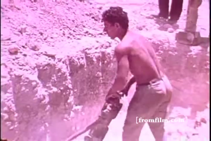 The still shows a young, shirtless male worker operating a jackhammer or similar tool in a dusty, excavated area. The scene is marked by a pinkish hue, characteristic of the film's color palette. The background is filled with loose soil and construction debris, indicating ongoing manual labor, likely related to a community or infrastructure project in an emergency context.