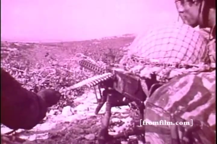 The still depicts a soldier in a camouflage uniform, positioned beside a mounted machine gun. The background features an expansive, rugged landscape. The image has a pinkish hue typical of older 16mm prints, suggesting a historical context related to military or emergency funding efforts in Israel. The soldier appears engaged, possibly in a tense situation, holding a hand toward the viewer.