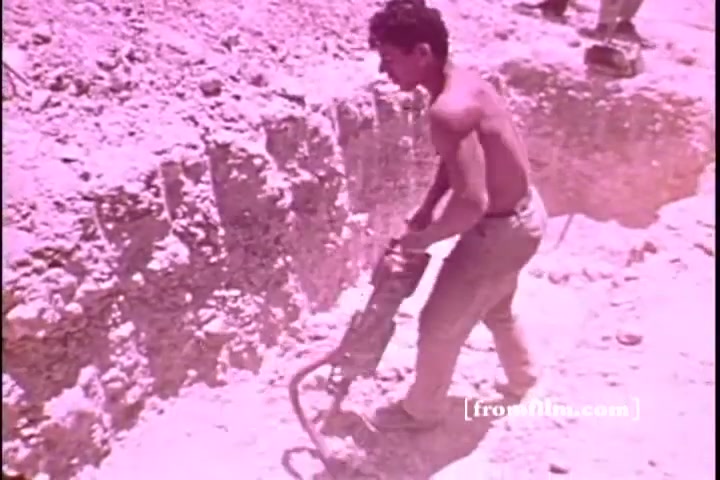 The still from the 'Israel emergency fund PSA' shows a young, shirtless man operating a jackhammer in a construction site. The environment appears dusty and rugged, suggesting an arduous labor context. The overall tone of the image is tinted, likely due to the film stock used, giving it a warm, pinkish hue. The scene conveys a sense of hard work and determination.