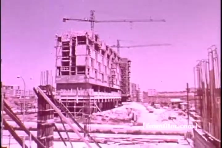 The still depicts a construction site with partially erected buildings and cranes, set against a pale pink backdrop typical of 16mm film. The foreground features scaffolding and construction materials, indicating active development in a developing area. The scene likely emphasizes growth and progress, relevant to the context of an emergency fund campaign for Israel.
