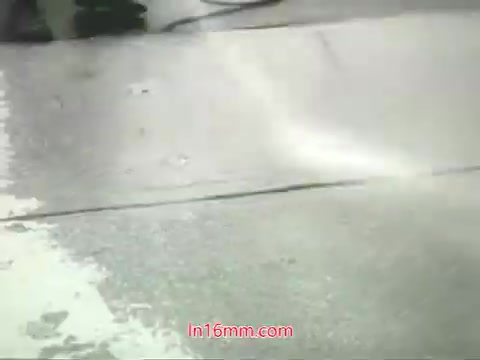 The still from the 16mm public service announcement features a city street scene, likely showing a sanitation or cleaning activity. The visuals may include water being sprayed or washed over the pavement, highlighting the challenges of urban pollution as described in Mason Adams' voiceover. The message emphasizes the inability to 'wash' the air we breathe, urging viewers to take action for cleaner air. The overall tone is one of awareness and responsibility regarding public health and environmental conditions in urban settings.