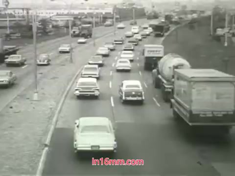 The 16mm still from the PSA depicts a busy urban highway in the 1960s. Numerous cars, predominantly vintage models, are seen traveling along the road which is flanked by signage and possibly industrial areas. The atmosphere appears overcast, hinting at pollution, which aligns with Mason Adams' voiceover regarding air quality issues in cities. The message emphasizes the inability to 'wash' the lungs clean from air pollutants, urging viewers to take action for cleaner air. The overall scene encapsulates the challenges of urban living and environmental health during that era.