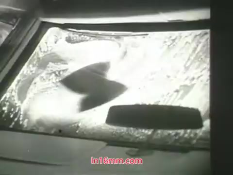 The still shows a windshield being washed, with soap suds and water splashing across the glass. It's a close-up shot that emphasizes the action of cleaning, likely symbolizing the effort to remove pollution and improve air quality. In the context of the PSA, it visually contrasts the idea of cleaning surfaces in a polluted city with the notion that lungs cannot be washed, underscoring the importance of addressing air quality issues. The imagery aligns with the message delivered by Mason Adams' voiceover, highlighting both the problem of pollution and a call to action for viewers.