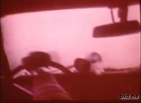 The still from 'Adventure: The Incredible Auto Race' captures a view from inside a car, showing the steering wheel and dashboard. The scene is tinted pink, suggesting an atmospheric or smoky environment that might indicate challenging road conditions. This perspective emphasizes the excitement and intensity of the road rally from London to Sydney, highlighting the driver's immersive experience during the race.