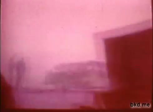 The still from 'Adventure: The Incredible Auto Race' (1970) features a hazy, overexposed look typical of 16mm film. The image captures an undefined scene, possibly an early rally moment, with two figures in the foreground and a vehicle blurred in the background. The atmosphere suggests a sense of adventure and excitement associated with the road rally from London to Sydney. The color palette leans towards pinkish tones, giving a nostalgic feel.