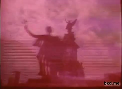 The still from the 'Adventure: The Incredible Auto Race promo (1970)' features a faded, pink-hued image of a statue against a cloudy sky. The statue, likely significant to the cinematic narrative, appears to be raising its arm, suggesting a sense of motion or announcement. The overall look reflects the vintage quality of a 16mm print, hinting at the excitement and adventure of the road rally from London to Sydney.