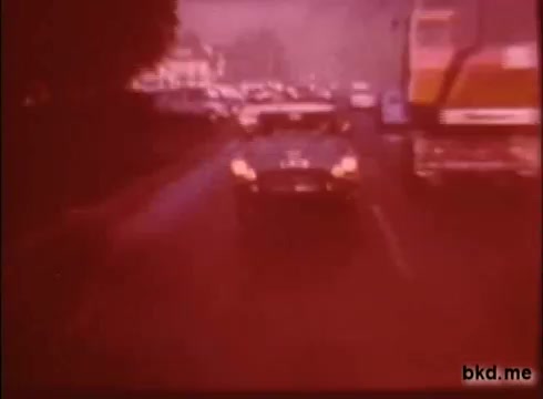 The still from 'Adventure: The Incredible Auto Race' (1970) showcases a bustling road scene, likely during the rally from London to Sydney. A vintage car is positioned prominently in the foreground, flanked by other vehicles, including a large truck. The image has a reddish tint typical of 16mm film prints, adding a nostalgic quality. The backdrop features blurred buildings, indicating a busy urban environment, while the overall atmosphere conveys a sense of motion and adventure.