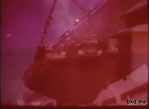 The still from 'Adventure: The Incredible Auto Race promo (1970)' shows a dramatic scene likely portraying the transportation of vehicles. The image has a reddish hue and appears to capture a moment involving a large ship or vessel, hinting at its role in the road rally from London to Sydney. The atmosphere suggests excitement and adventure, typical of auto racing promos.