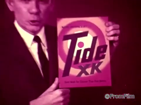 The still features a man in a suit presenting a box of Tide XK laundry detergent, emphasizing its stain-removing capabilities. The commercial implies that it can effectively tackle tough stains like blood and ketchup, showcasing a breakthrough in cleaning technology. The context suggests a humorous or dramatic situation involving a guest towel and the high expectations surrounding stain removal.