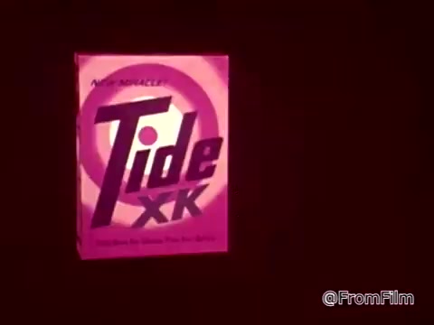 The 16mm still features a vibrant pink box of 'Tide XK' laundry detergent, prominently displaying the product name in bold lettering. The background has a deep red hue, enhancing the product's visibility. The commercial context implies a humorous scenario involving blood and ketchup stains, emphasizing the effectiveness of Tide XK in tackling tough stains. A voiceover highlights that traditional stain barriers have been broken, showcasing the detergent as a revolutionary cleaning solution.