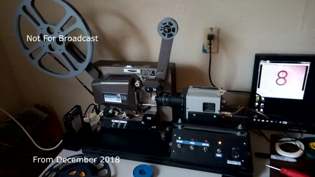 The still captures a film transfer setup from December 2018, featuring a projector connected to a video capture device. The projector is displaying a reel of 16mm film, indicating the transfer process from film to video. A computer monitor nearby likely shows the capturing interface or playback, and there are control buttons and tools on the workstation. The labeling 'Not For Broadcast' suggests the material is intended for private use rather than public airing, and the mention of audio sync issues implies technical challenges faced during the transfer.