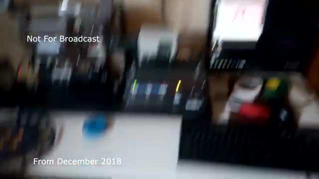 The still captures a workspace focused on the transfer of a film to video, specifically for a TV commercial from December 2018. The setup includes equipment like film projectors and video playback devices, with various buttons and lights indicating operation. There are hints of clutter, suggesting an active editing environment, while a few pieces of technology hint at the audio sync issues you aim to address. Overall, the image conveys a behind-the-scenes look at the transfer process for archived footage.