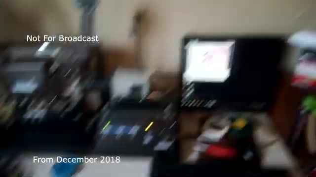 The still showcases a behind-the-scenes look at the process of transferring a 16mm film commercial to video. It appears cluttered with equipment, including a film projector and a video monitor. There are some visible indicators of activity, such as control lights, suggesting the transfer process is in progress. The image includes text that states 'Not For Broadcast' and 'From December 2018,' indicating its archival nature and non-broadcast purpose. However, the blurriness might reflect technical issues, possibly including the aforementioned audio sync problems.