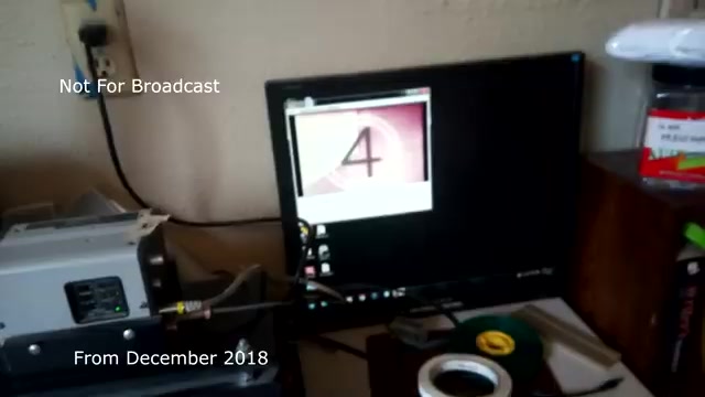 The still from 'December 2018 transfer talk: Chase and Sandborn Commercial' shows a workstation where a 16mm film is being prepared for transfer to video. In the foreground, there's a film projector, and a computer monitor displays a frame number, indicating that the transfer process is in progress. The setup has various items like tape and other equipment typical for film editing and transfer work. The text on the image states that it is 'Not For Broadcast' and marks it as from December 2018. The mention of prior audio sync issues suggests that this clip addresses technical challenges faced during the transfer process.