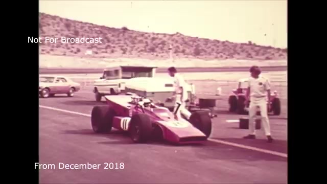 The still from the 'December 2018 transfer talk - Chase and Sanborn Commercial' showcases a vintage racing scene captured on 16mm film. In the image, a classic race car is positioned on a track, surrounded by crew members in white uniforms. Nearby, several vehicles are visible, indicating a bustling pit area. The image bears a distinct retro aesthetic, highlighting the era’s racing culture. The accompanying text, “Not For Broadcast” and the date, emphasizes its archival nature and potential audio sync issues that were later addressed. This snapshot reflects the technical transition from film to video for the commercial.