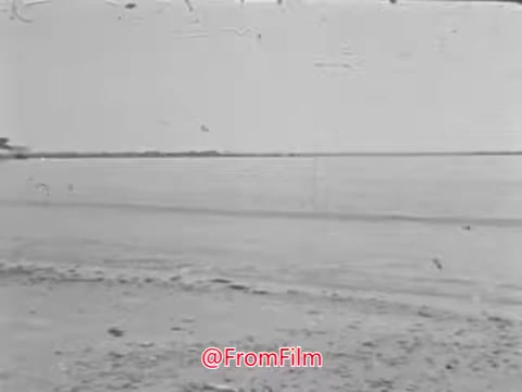 The still features a serene beach scene with gentle waves lapping at the shore, captured in a vintage 16mm film style. The atmosphere is tranquil, evoking a sense of timelessness in line with the voice-over reflecting on the enduring nature of the sea. The image likely aims to inspire a sense of belonging and adventure associated with naval service.