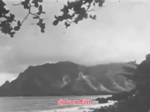 The still from the 'US Navy Recruiting PSA - 1960s' features a tranquil coastal scene. The image captures a rocky shoreline with waves gently washing ashore, framed by overhanging leaves and a backdrop of a dramatic mountain landscape under a moody sky. The atmosphere evokes a sense of timelessness, aligning with the voice-over's poetic reflection on the sea's enduring presence. This scene exemplifies the allure of the Navy and the call to adventure.