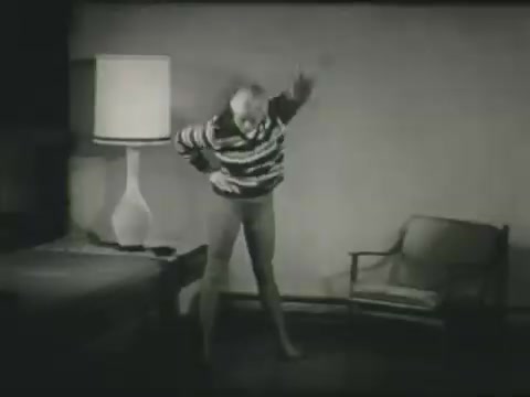 The still features a person in a striped sweater and light-colored pants, performing an exaggerated motion, likely meant to illustrate a skiing safety message. The background includes a bedside lamp and a chair, suggesting an indoor setting. The image likely aims to convey a humorous or instructional tone regarding skiing precautions.