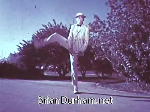 The still from the 1968 Red Rose Tea commercial features a man dressed in a light striped suit and hat, performing a playful dance move with one leg raised high. The background includes trees, suggesting an outdoor setting. The overall atmosphere appears cheerful and whimsical, typical of mid-20th century advertisements aimed at promoting the product.