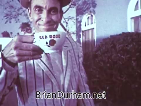 The still from the 1968 'Another Red Rose Tea' commercial features a man in a striped suit, grinning and holding a cup that prominently displays 'Red Rose.' The backdrop includes a well-maintained garden, adding a cheerful and inviting atmosphere to the scene. The overall feel is nostalgic and reflects the advertising style of the era, aiming to promote the brand in a friendly manner.