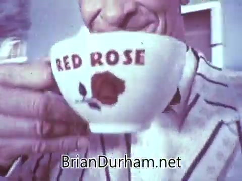 The still from the 1968 'Another Red Rose Tea' commercial features a close-up of a man happily sipping from a cup adorned with the words 'RED ROSE' and a rose graphic. He is wearing a striped shirt, suggesting a casual and cheerful atmosphere. The visual style reflects the era's vibrant color palette and advertising aesthetics. The context hints at a promotional piece that captures the essence of enjoying tea.