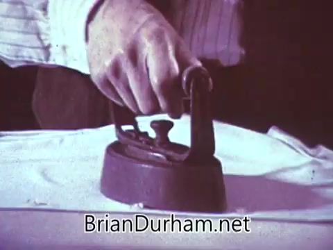 The still from the 'Another Red Rose Tea' commercial features a close-up shot of a hand gripping a vintage iron, pressing it down onto a piece of fabric. The scene suggests a domestic setting and emphasizes traditional household tasks, typical of commercials from the 1960s. The color palette appears muted, characteristic of the era's filming techniques. The overall context hints at a nostalgic representation of home life, likely linking the act of ironing to comfort and quality, paralleling the experience of enjoying Red Rose Tea.