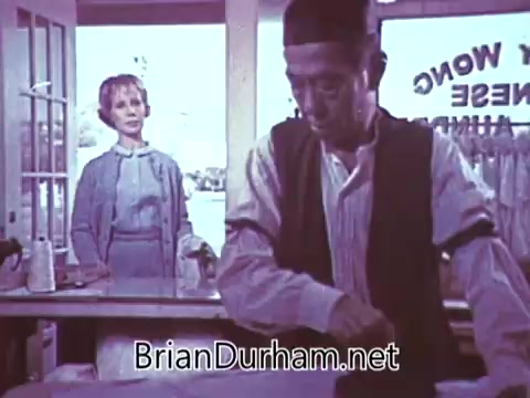 The still from the 1968 'Another Red Rose Tea' commercial features a scene in a tea shop. A man, dressed in traditional clothing, is preparing something at a counter, focused on his work. In the background, a woman watches intently, perhaps displaying curiosity or interest in the tea preparation. The setting appears bright, suggesting an inviting atmosphere typical of tea shops. The composition highlights both the man’s actions and the woman’s engagement, emphasizing the theme of tea culture.