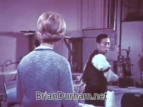 The still from the 1968 'Another Red Rose Tea' commercial shows a scene featuring two characters. One character, likely a woman, has her back to the camera, wearing a light-colored sweater, while the other, a man, is facing her and appears to be in motion, possibly engaged in some activity related to tea preparation. The setting looks formal or professional, suggesting it might be set in a kitchen or studio environment. The color palette is soft, typical of mid-20th century commercials.