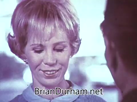 The still from the 1968 'Another Red Rose Tea' commercial features a woman with short, light-colored hair, smiling and appearing to engage in conversation. The background is blurred, creating a soft focus effect that emphasizes her expression. The overall tone suggests a warm and inviting atmosphere, typical of advertising from that era, aimed at promoting the tea brand's approachable and friendly image.