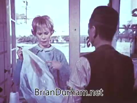 The still from the 1968 'Another Red Rose Tea' commercial features a woman with short, light hair, appearing surprised or intrigued as she examines an object from a bag. Opposite her is a man in historical attire, likely portraying a character relevant to the tea theme. The setting looks like a shop or market, giving off a vintage feel consistent with the time period. The overall tone suggests a lighthearted or playful interaction centered around tea.