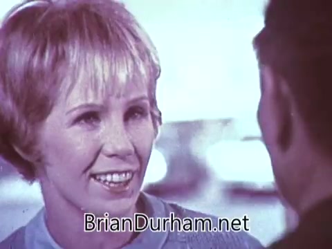 The still from the 1968 'Another Red Rose Tea' commercial features a woman with short, light blonde hair and a friendly smile. She appears to be engaged in conversation, likely promoting the tea. The background is softly blurred, emphasizing her expression and the interaction. The visual style reflects the era's playful and vibrant advertisements, capturing the essence of mid-century advertising aesthetics.