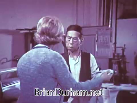 The still features two individuals engaged in a conversation, likely in a domestic or casual setting. One person appears to be an older male, while the other is a female with short hair. They seem to be discussing something with an air of curiosity or intrigue. The color palette has a vintage quality typical of the era, possibly contributing to a nostalgic ambiance. The focus of the discussion may relate to Red Rose Tea, given the context.