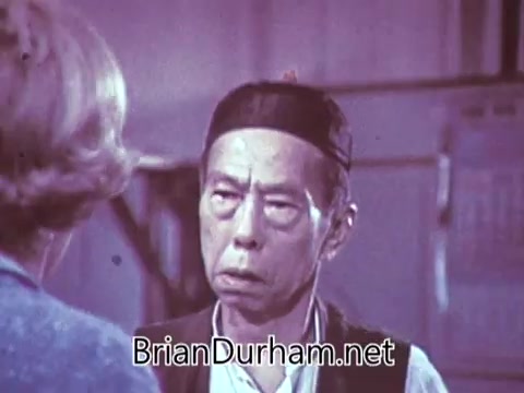 The still from the 1968 Red Rose Tea commercial features a man with an intense expression, wearing a traditional outfit and a cap. He appears to be engaged in conversation with a woman, who is partially visible on the left side of the frame. The overall color palette has a vintage feel, characteristic of the era's advertising style. The setting seems to suggest a cultural or thematic context surrounding tea preparation or enjoyment.