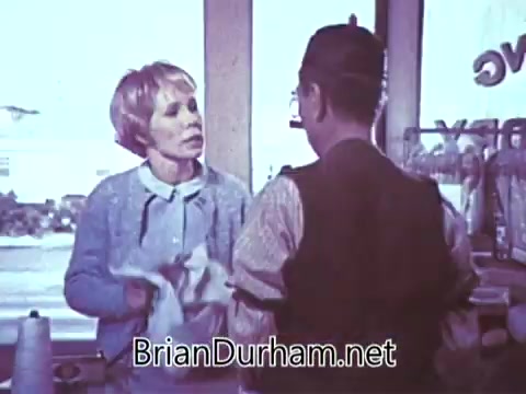 The still shows a woman and a man engaged in conversation, likely in a café or shop setting. The woman appears to be holding a towel, suggesting she may have just finished cleaning or serving. The environment is bright and showcases a casual interaction, typical of advertising from the late 1960s. The focus on the characters hints at a personal connection, likely emphasizing the warmth associated with Red Rose Tea.