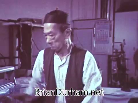 The still from the 1968 'Another Red Rose Tea' commercial features a man in a black cap and vest, engaged in a task, presumably related to tea preparation. The setting appears to be a workshop or kitchen, with various equipment visible in the background. The overall tone of the image is nostalgic, characteristic of the era's advertising style, aiming to evoke warmth and authenticity in promoting the tea brand.