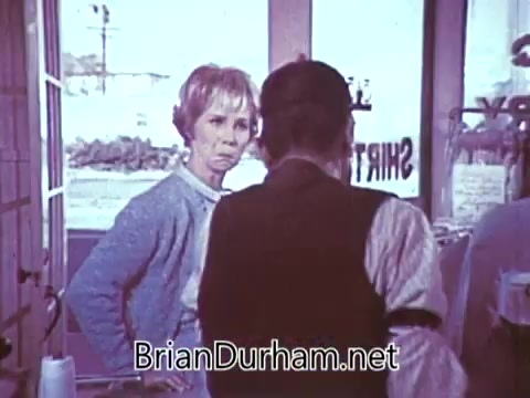 The still from the 1968 'Another Red Rose Tea' commercial features a woman with a concerned expression, looking towards a person whose back is facing the camera. The setting appears to be a small shop or café, with a window displaying a view outside. The overall mood suggests an interaction about a product, likely emphasizing the qualities of Red Rose Tea. The vintage aesthetic is characteristic of the era.