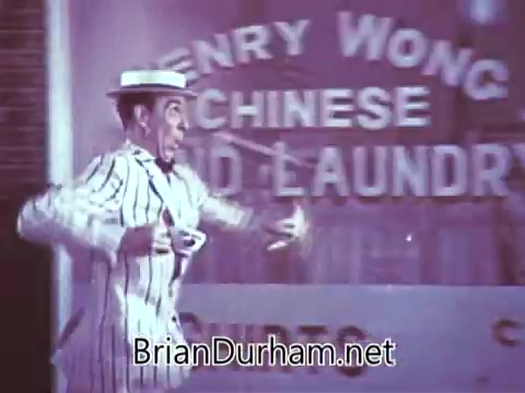 The 16mm still from the 1968 'Another Red Rose Tea' commercial features a man in a classic striped suit and hat, energetically promoting Red Rose Tea. He appears enthusiastic, possibly engaging with the camera or audience. The background includes a storefront labeled 'Henry Wong Chinese Laundry,' adding a whimsical or light-hearted context to the scene. The overall tone suggests a playful and vintage advertising style typical of that era.